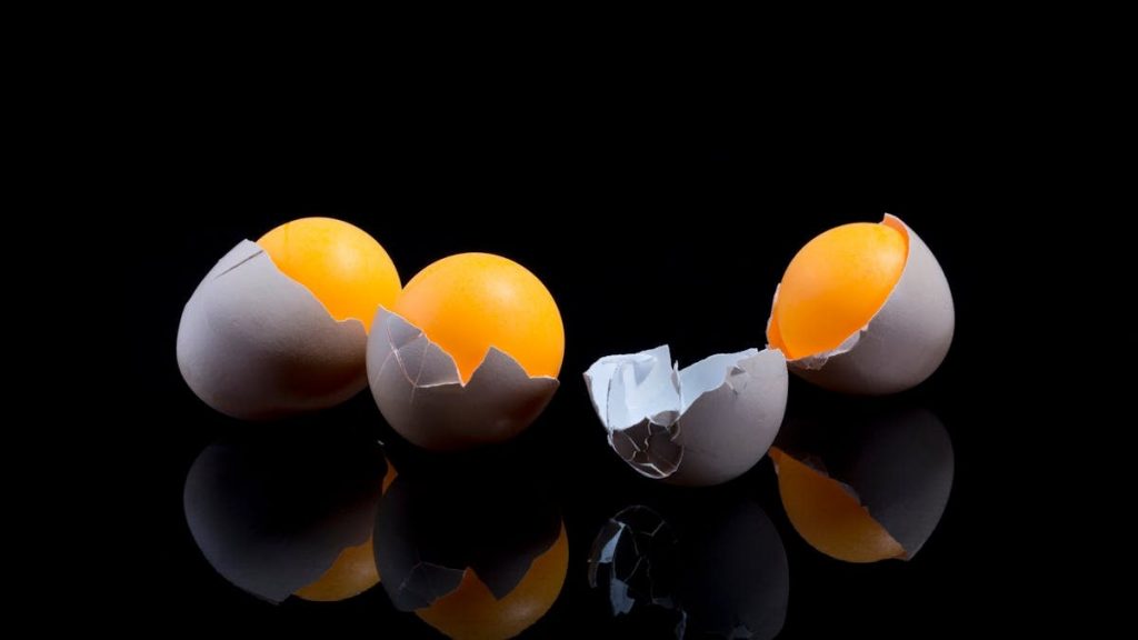 Two cracked eggs with bright yolks on a reflective black surface, showcasing culinary ingredients.