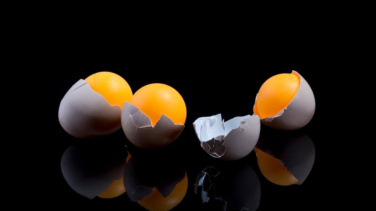 Two cracked eggs with bright yolks on a reflective black surface, showcasing culinary ingredients.