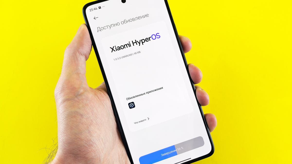 Hand holding smartphone showing Xiaomi HyperOS update screen on yellow background.
